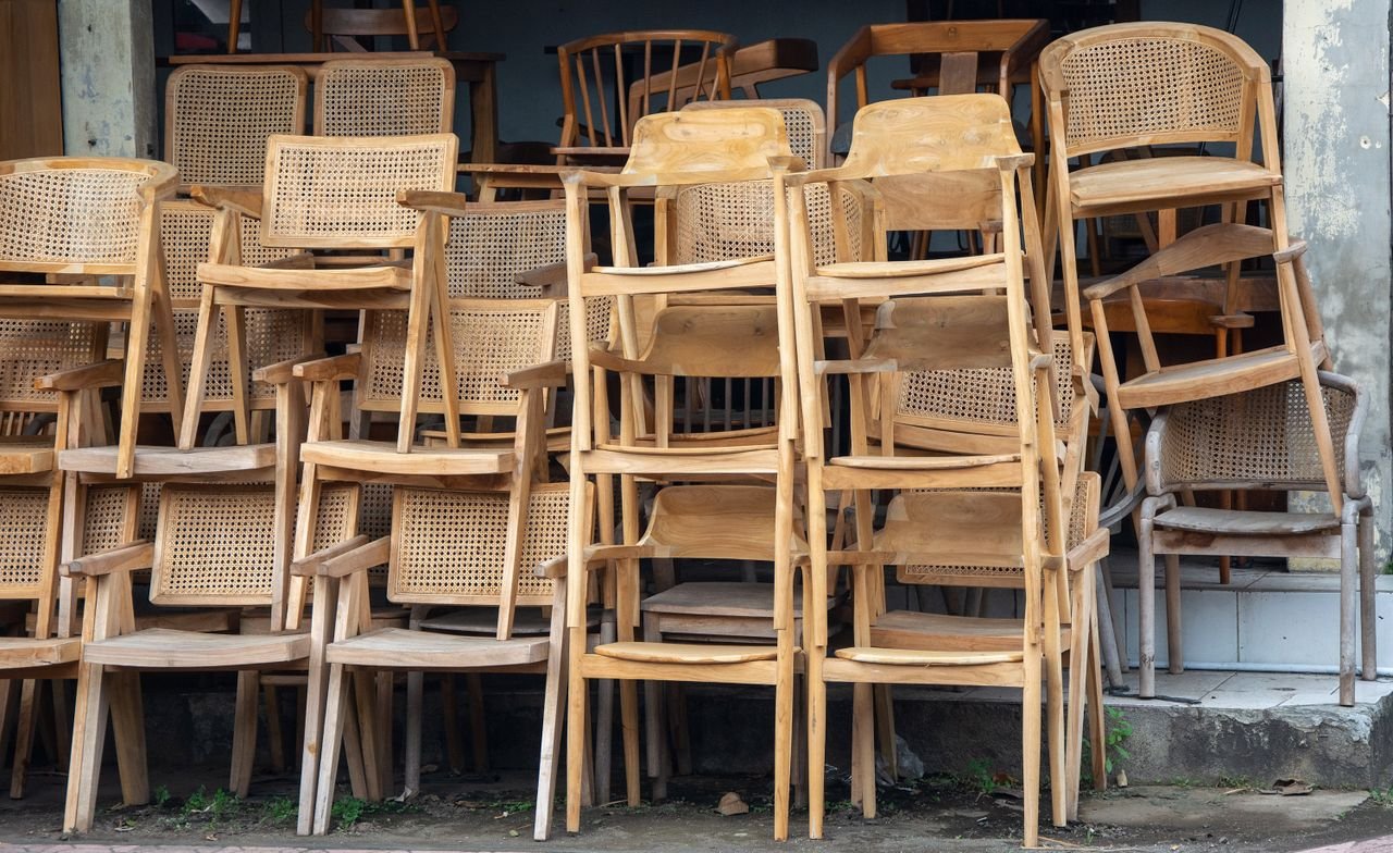 An array of stacked wooden and wicker chairs leaning against a weathered urban wall.