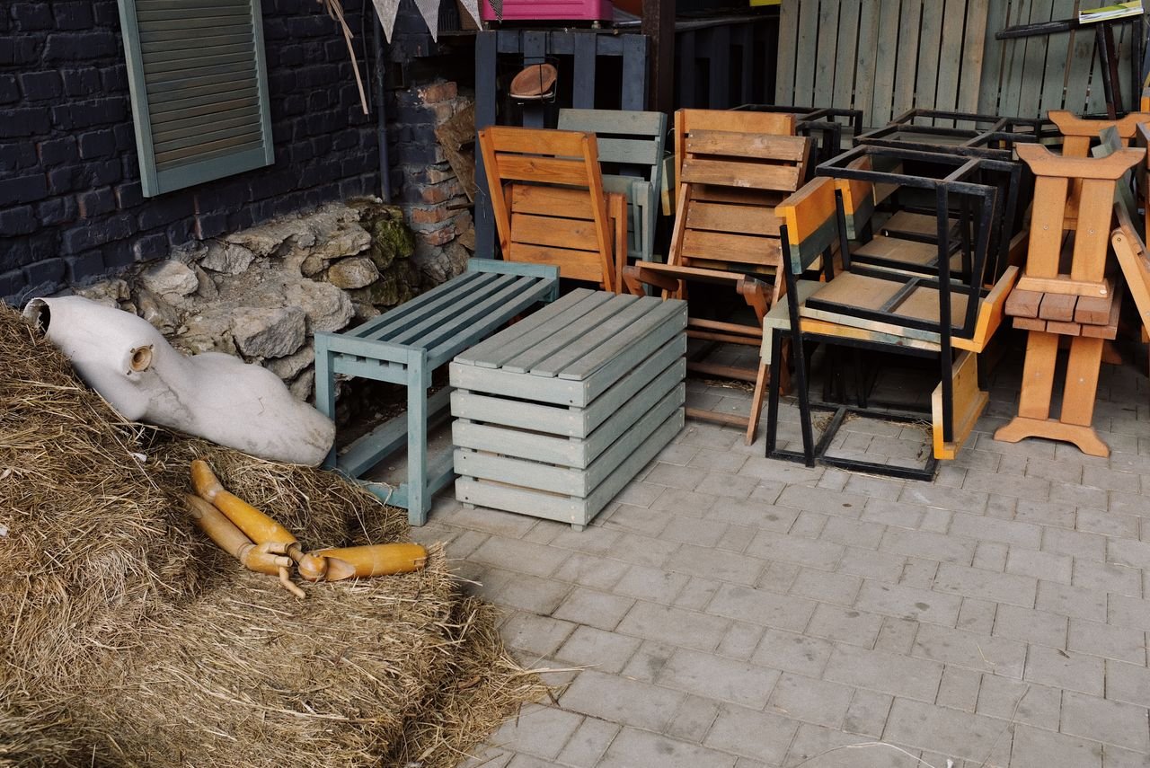Stacked wooden furniture and straw bales outdoors against a stone wall, creating a rustic scene.